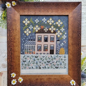 May include: A cross-stitch embroidery depicting a house with a garden. The house is pink with white trim and has a green roof. The garden is filled with flowers in shades of blue, pink, and white. There are also a few birds and a bee in the garden.