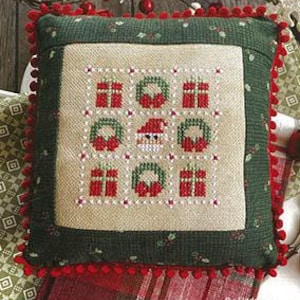 May include: A square decorative pillow with a green border and a central embroidered design. The design features red gift boxes, wreaths, and a Santa Claus face. The pillow is trimmed with red pom-poms and has the text "December Nine Patch" at the top.