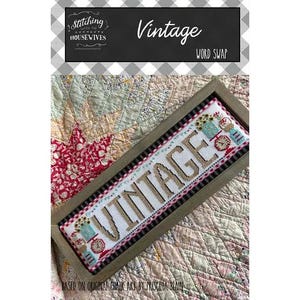 May include: A framed cross-stitch piece with the word "VINTAGE" in tan thread. The piece is bordered with a black and white pattern and decorative elements. The background is a patterned quilt.