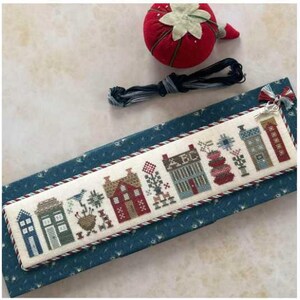 May include: A cross-stitched bookmark featuring a row of colorful houses and floral designs. The bookmark is bordered with a twisted red and white cord and rests on a blue patterned surface. A red tomato pin cushion and black thread are also present.