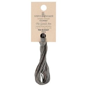 Puede incluir: Una madeja de hilo de bordar gris con reflejos marrones y azules. El hilo está etiquetado con "Simply Shaker Sampler Threads Colorfast The Gentle Art Counted Thread Embroidery PO Box 670 New Albany, Ohio 43054 Tin Bucket 7024"