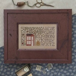 May include: Cross-stitch pattern featuring a red house with a window and a beehive. The alphabet is stitched in black thread below the house and beehive. The pattern is framed in a dark wood frame.