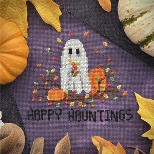 Könnte beinhalten: Ein Kreuzstich-Design mit einem weißen Geist, Kürbissen und Herbstblättern auf lila Hintergrund. Der Text "HAPPY HAUNTINGS" ist darunter gestickt. Gelbe und braune Blätter und ein Kürbis umgeben das Design, passend zu Halloween.