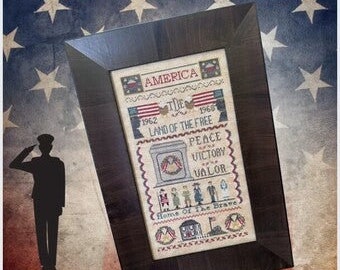May include: Framed cross-stitch art with a patriotic theme. The artwork features the text "America," "Land of the Free," and "Peace, Victory, Valor." The frame is dark brown, and the background has a flag design with stars.