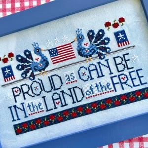 May include: Cross-stitch artwork in a blue frame. The design features two blue birds holding an American flag, with the text "PROUD AS CAN BE IN THE LAND OF THE FREE." The artwork includes red, white, and blue accents, and floral details.