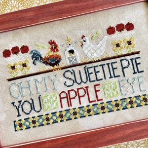 Puede incluir: Obra de arte de punto de cruz en un marco de madera. El diseño presenta un gallo, una gallina, manzanas y el texto "OH MY SWEETIE PIE YOU APPLE OF MY EYE". La paleta de colores incluye rojo, azul, verde, amarillo y blanco.