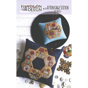 Peut inclure: Un motif de point de couture vintage avec un design hexie. L'image montre deux coussins décoratifs avec des détails brodés, dont des ciseaux et des motifs floraux. Les coussins sont dans les tons de bleu, beige et rouge. Le texte "HANDS ON DESIGN" et "A VINTAGE STITCH" est visible.