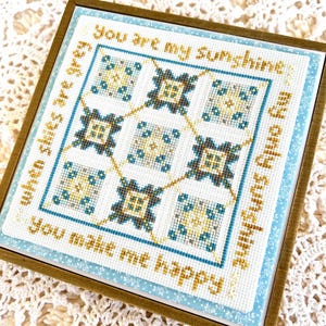 May include: A framed cross-stitch with a blue and gold geometric pattern and the text "You are my sunshine, my only sunshine, You make me happy" in a white font.
