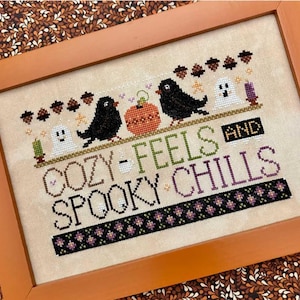 May include: Cross-stitch artwork in a wooden frame. The design features black birds, a pumpkin, and ghosts. Text reads "Cozy Feels and Spooky Chills." The color palette includes black, orange, green, and purple. The background is a light beige.
