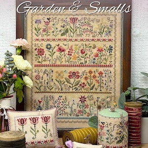 Peut inclure: Une broderie au point de croix encadrée intitulée "Spring Alphabet Garden & Smalls" avec des motifs floraux et alphabétiques. Un coussin et un coussin à épingles avec broderie florale sont au premier plan, avec des bobines de fil et un récipient décoratif.