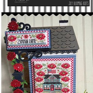 May include: A decorative wooden house-shaped sign featuring cross-stitch designs. The sign includes the text "Zinnia Lane" and a house with an American flag. Red flowers and a gingham star add to the patriotic theme.