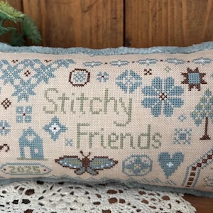 May include: A rectangular, beige pillow with a light blue fringe. The pillow features cross-stitch embroidery with the words "Stitchy Friends" and various floral and geometric patterns in shades of blue and brown. A butterfly and a house are also embroidered.