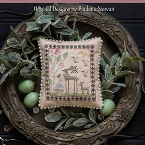 May include: A cross-stitch sampler featuring a deer, birds, and flowers in a brown frame. The sampler is on a vintage metal tray with green leaves and eggs. The text "Spring Beginnings" is at the bottom of the image.