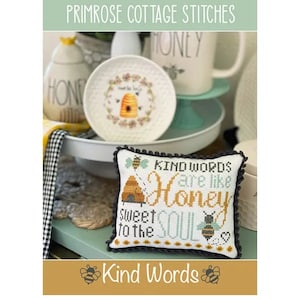 May include: A white pillow with black trim features a cross-stitch design of a beehive and the text "Kind Words are like Honey Sweet to the Soul". The pillow is on a table with a white plate and a honey-themed pitcher.