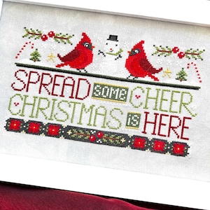 May include: Cross-stitch artwork in a white frame. The design features two red cardinals, a snowman, and the text "SPREAD SOME CHEER CHRISTMAS IS HERE" in various colors. Festive holiday decor.