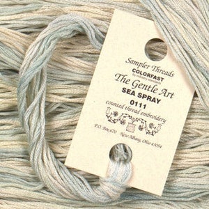 May include: A skein of light blue, beige, and white embroidery floss with a label that reads "Sampler Threads Colorfast The Gentle Art Sea Spray 0111 counted thread embroidery P.O. Box 670 New Albany Ohio 43054"