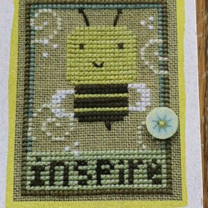 May include: Cross-stitch design featuring a smiling bee with black and yellow stripes, surrounded by white accents. The word "INSPIRE" is stitched below the bee. The design is framed in a yellow border and includes a small, light blue button.