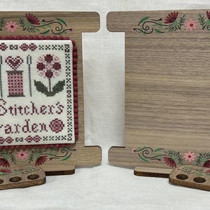 Op de afbeelding: Houten kruissteek displaystandaard met een gestikt ontwerp met de tekst "A Stitcher's Garden". De standaard heeft een lichtbruine afwerking met bloemaccenten in roze, groen en bordeauxrood. Inclusief een kleine houten draadhouder.