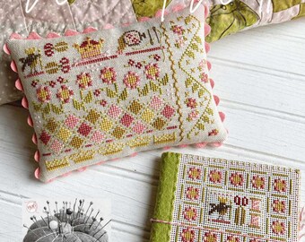 Op de afbeelding: Een kussen met borduurwerk in kruissteek met een bloemenmotief en de tekst "Wear it well". Het kussen ligt op een wit houten oppervlak met een groen en roze quilt op de achtergrond. Er is ook een klein groen en wit kruissteekboekje en een naaldenkussen met de tekst "Pins & Needles".