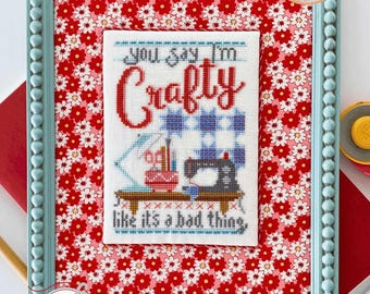 Op de afbeelding: Een ingelijst kruissteekontwerp met een blauwe en witte achtergrond en rode tekst die "You say I'm Crafty like it's a bad thing." luidt. De lijst is lichtblauw met een wit bloemenpatroon op de passe-partout.