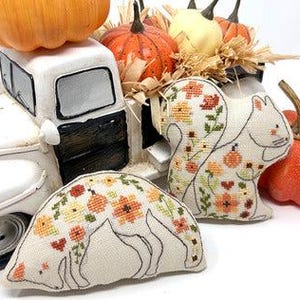 May include: Two white fabric squirrel-shaped pillows with embroidered floral and pumpkin designs. The pillows are sitting on a white toy truck bed filled with hay and pumpkins.