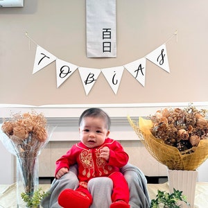 Peut inclure: Un bébé garçon portant une tenue traditionnelle chinoise rouge avec des broderies dorées est assis sur une couverture grise. Le bébé est entouré de décorations, dont une bannière qui dit "Tobias" et une bannière blanche avec des caractères chinois.
