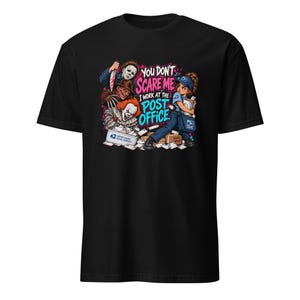 Könnte beinhalten: Ein schwarzes T-Shirt mit einem Grafikdruck, der eine Postkraft umgeben von Horrorfilmfiguren zeigt. Der Text lautet: "YOU DON'T SCARE ME I WORK AT THE POST OFFICE." Das Design enthält Post, ein USPS-Logo und farbenfrohe Spritzer-Akzente.