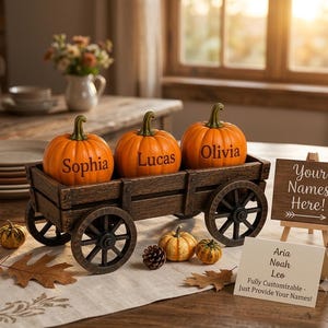 May include: A dark brown wooden wagon containing three orange pumpkins with the names Sophia, Lucas, and Olivia engraved on them. A small wooden sign on an easel displays the text Your Names Here! and a second card lists Aria, Noah, Leo.