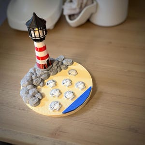 May include: A decorative lighthouse figurine with a red and white striped tower and a black top. The lighthouse sits on a circular base with seashells and a blue wave design. The base is approximately 4 inches in diameter.