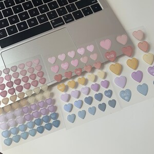May include: Heart-shaped stickers in various pastel colors, including pink, yellow, purple, and blue. Some stickers have phrases like "LOVE," "KISS," and "CALL ME." The stickers are arranged on clear sheets.