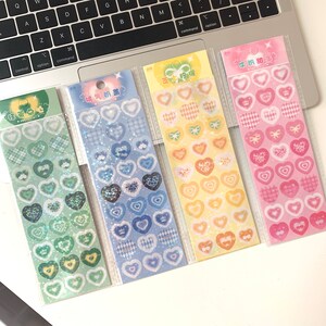 May include: Four sticker sheets with heart-shaped designs in various colors. The stickers feature patterns like plaid, bows, and crowns. The color palette includes green, blue, yellow, and pink, with some stickers having a glittery effect. The sheets are packaged in clear plastic.