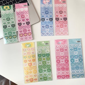 May include: A collection of heart-shaped stickers in various colors and patterns. The stickers are arranged in vertical strips and packaged in clear plastic. Designs include gingham, bows, and floral motifs. The color palette includes shades of green, pink, blue, and yellow.