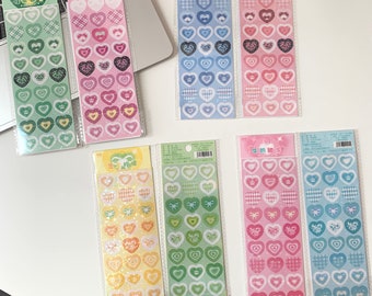 May include: A collection of heart-shaped stickers in various colors and patterns. The stickers are arranged in vertical strips and packaged in clear plastic. Designs include gingham, bows, and floral motifs. The color palette includes shades of green, pink, blue, and yellow.