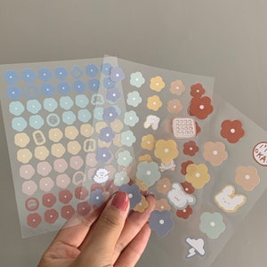 May include: Sheets of colorful flower stickers in pastel shades of blue, yellow, and brown. Some stickers have small illustrations of animals and food. The stickers are held by a hand.
