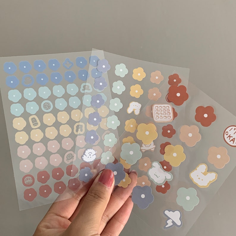 May include: Sheets of colorful flower stickers in pastel shades of blue, yellow, and brown. Some stickers have small illustrations of animals and food. The stickers are held by a hand.