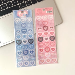 May include: Two sticker sheets with heart-shaped designs. One sheet features blue hearts with various patterns, including plaid and leopard print. The other sheet has pink hearts with similar patterns. The stickers are packaged in clear plastic.