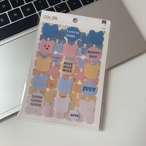 May include: A clear plastic package of decorative stickers featuring bear shapes and circular designs. The stickers are in pastel colors, with text like "I will be happy", "Nice", and "Just". The package is on a laptop and keyboard.