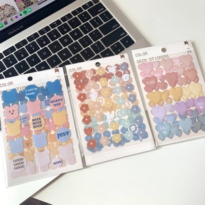 May include: Three packs of decorative stickers with various designs. One pack features bear shapes and text like "Nice Day". Another has flower shapes in pastel colors. The third pack contains heart shapes with phrases like "Kiss" and "Hug Me".