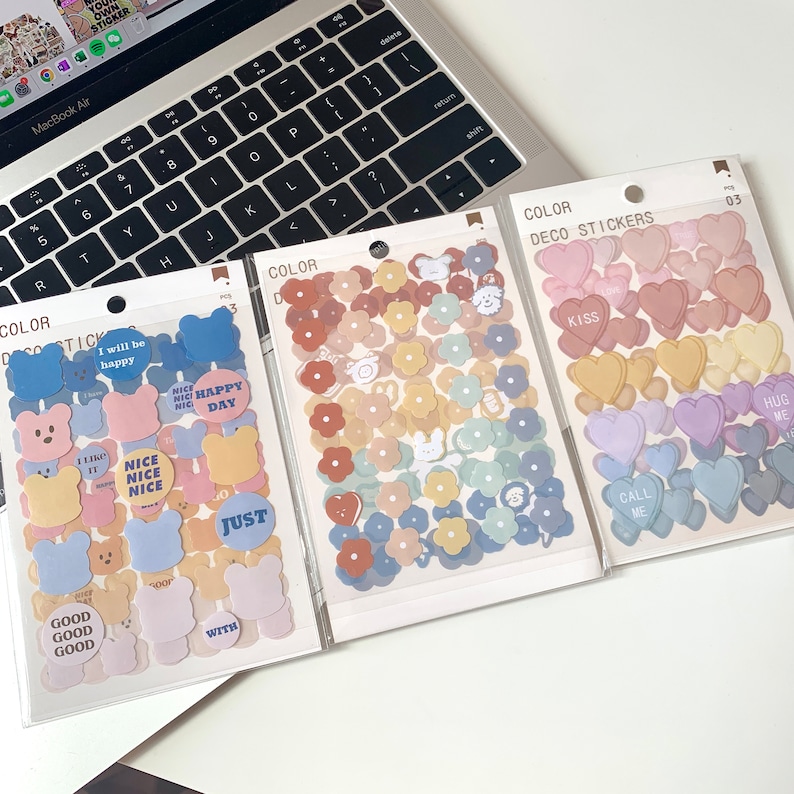 May include: Three packs of decorative stickers with various designs. One pack features bear shapes and text like "Nice Day". Another has flower shapes in pastel colors. The third pack contains heart shapes with phrases like "Kiss" and "Hug Me".