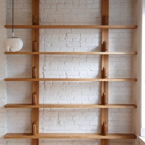 May include: A white brick wall with a wooden shelf unit. The shelf unit has five shelves and is made of natural wood. The shelves are supported by two vertical wooden posts.