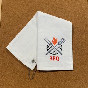 May include: White kitchen towel with a red embroidered BBQ logo featuring a crossed fork and spatula with a flame in the center.