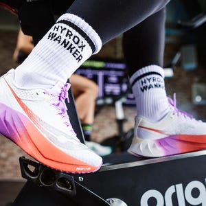 May include: White athletic sneakers with purple and orange gradient soles and purple laces, paired with white crew socks featuring black text that reads HYROX WANKER, worn while using a black exercise machine with the word origin visible.