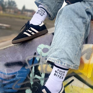 May include: White socks with the text "THIS BITCH BITES" in black, worn with black trainers featuring white stripes and a brown sole. The person is wearing light blue jeans. The background is a graffiti-covered wall.