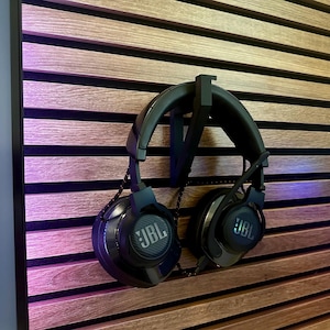 May include: Black JBL headphones suspended from a black holder on a wooden slat wall. The headphones display the JBL logo on the earcups. The wall is constructed of horizontal wooden slats.