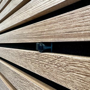 Puede incluir: Primer plano de listones de madera con un acabado texturizado marrón claro. Los listones están dispuestos en líneas paralelas, creando un patrón. Un pequeño objeto cilíndrico negro es visible entre los listones. El fondo es oscuro.