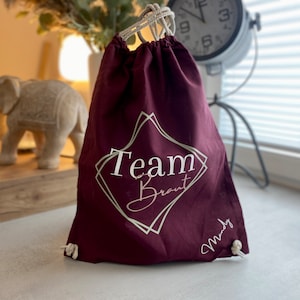 Pode incluir: Uma bolsa de cordão bordô com o texto "Team Braut" em um design em forma de diamante. A bolsa também tem o nome "Mandy" escrito. Possui um fecho de cordão branco.