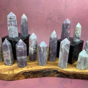 May include: A collection of 14 purple and green lepidolite crystal points arranged on a wooden display stand. The points are various sizes and shapes, with some having a more pronounced point than others.