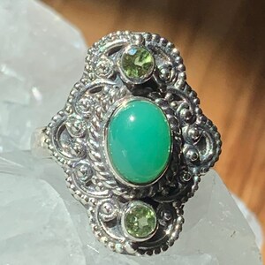 May include: A silver ring with an ornate design featuring a large oval green gemstone and two smaller green gemstones.