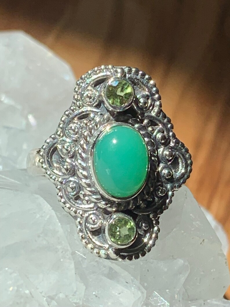 May include: A silver ring with an ornate design featuring a large oval green gemstone and two smaller green gemstones.