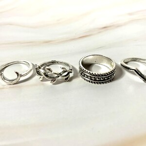 May include: Four silver rings of varying designs are displayed. One ring has a crescent shape, another features a leaf design, a third has a beaded band, and the last is a simple V-shape. The rings are on a light-colored surface.